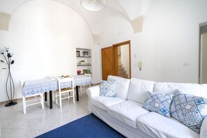 A living room with a white sofa, a dining table with a blue tablecloth, and a staircase leading to another level.