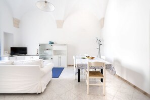 A modern living room with a white sofa, a dining table with a patterned tablecloth, and a television on a cabinet.