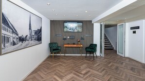 A reception area with a check-in/check-out desk, two green chairs, a large framed picture, and a herringbone patterned floor. - Hotel Das Q Spaichingen (Spaichingen)