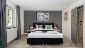A modern bedroom with a large bed, bedside tables, a framed artwork, a radiator, and a door to a shower area. - Hotel Das Q Spaichingen (Spaichingen)