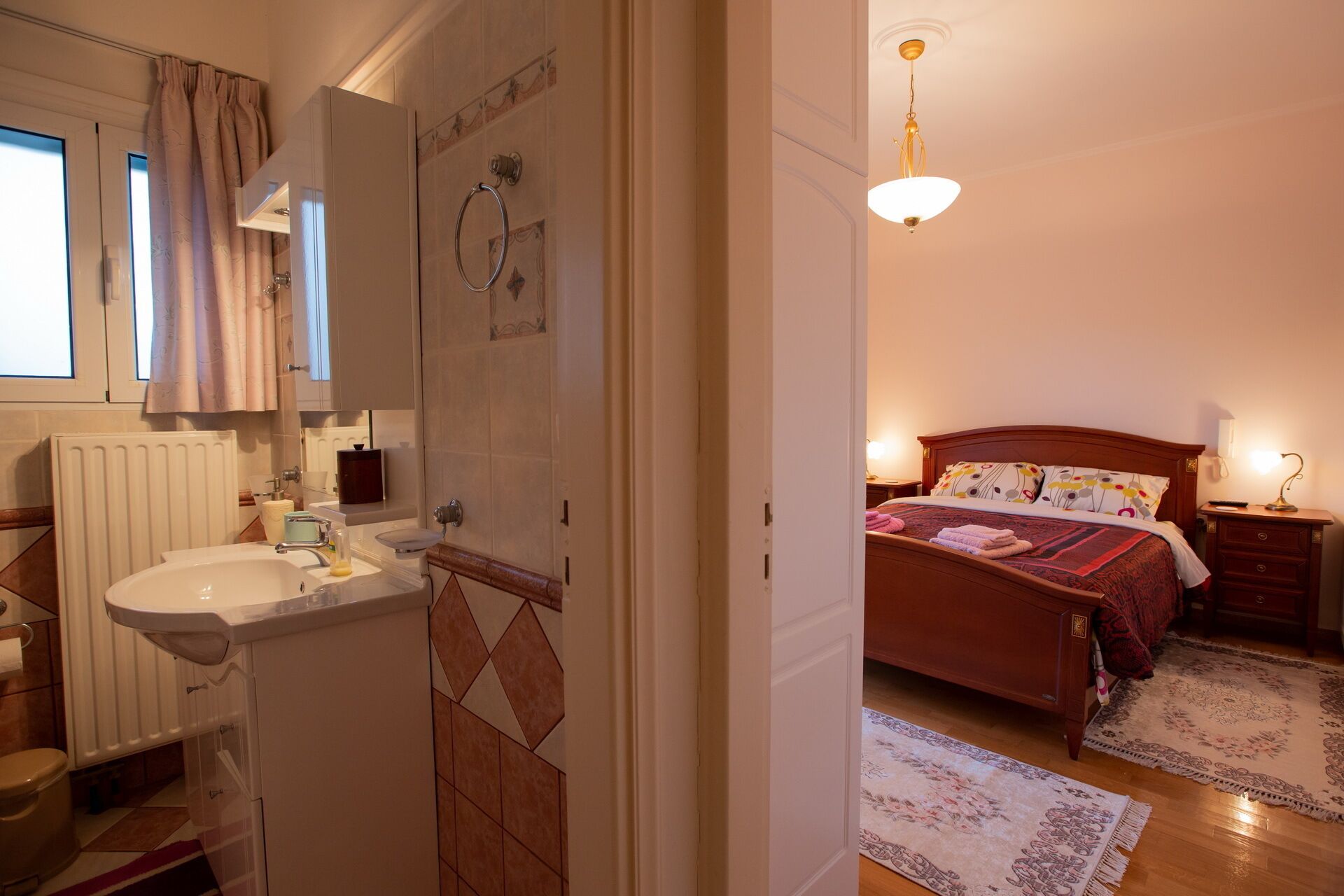 A bathroom with a sink, toilet, and shower area. The bedroom features a bed, nightstand, and a hanging light fixture.