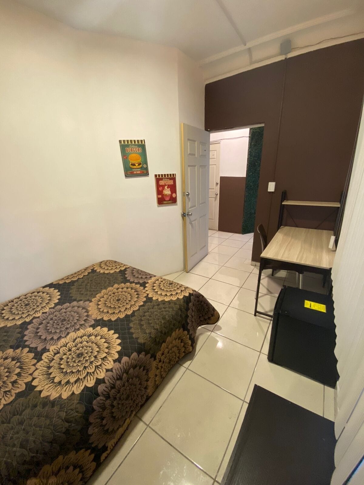 Una habitación pequeña y limpia con cama, una mesa y una silla. Hay dos carteles en la pared y una puerta que da a otra habitación.