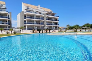 A modern hotel with a swimming pool, multiple balconies, and a clear blue sky.