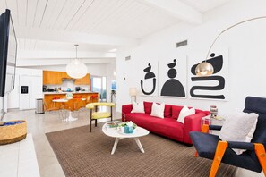 A modern living room with a red sofa, a blue armchair, and a coffee table. There is a kitchen in the background with wooden cabinets and a dining area.