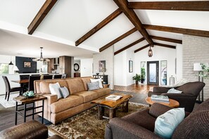 A spacious living room with a high ceiling, wooden beams, a large sofa, a coffee table, and a dining area with a kitchen in the background.