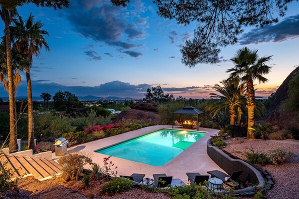 A swimming pool with lounge chairs and a gazebo, surrounded by palm trees and a view of distant mountains.