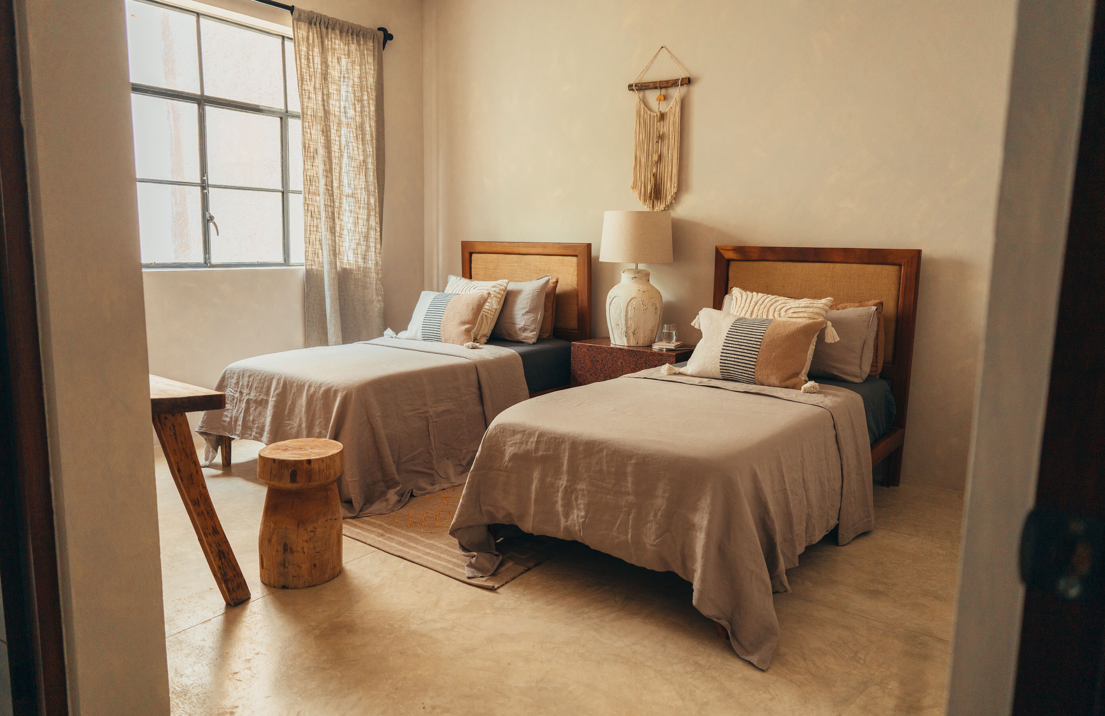 A room with two beds, a wooden table, a lamp, and a macrame wall hanging.