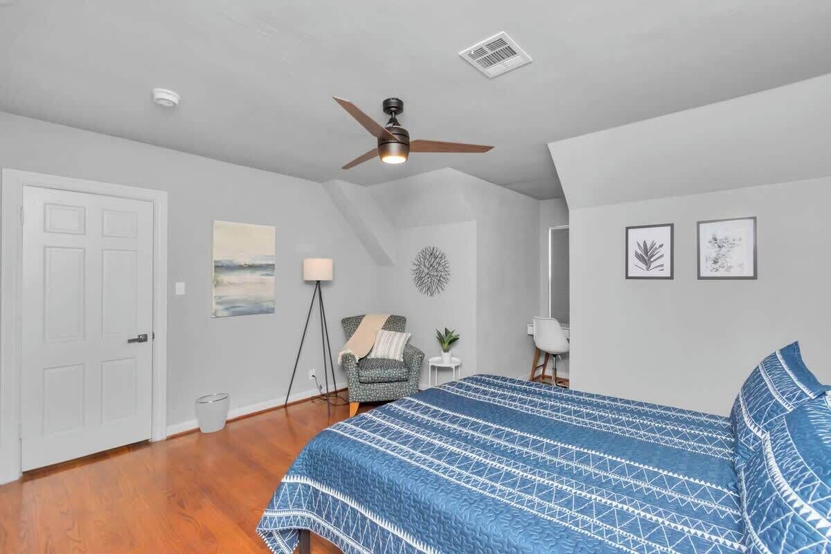 A bedroom with a bed, a ceiling fan, a chair, a lamp, and framed artwork on the wall.