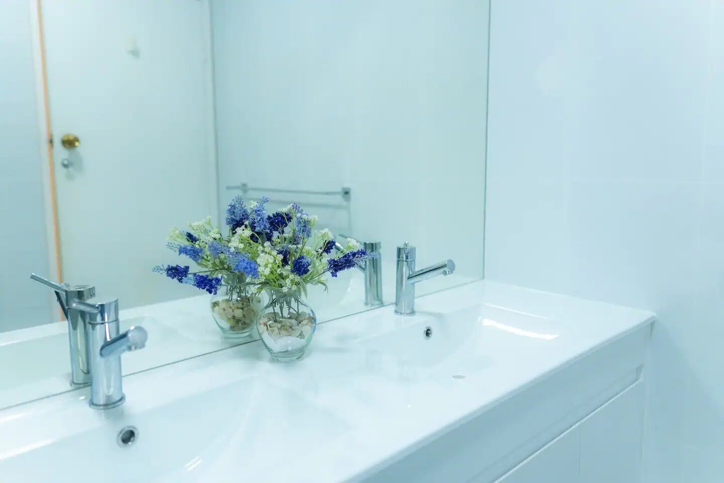 Un lavamanos blanco con dos jarrones de vidrio con flores y un espejo encima.