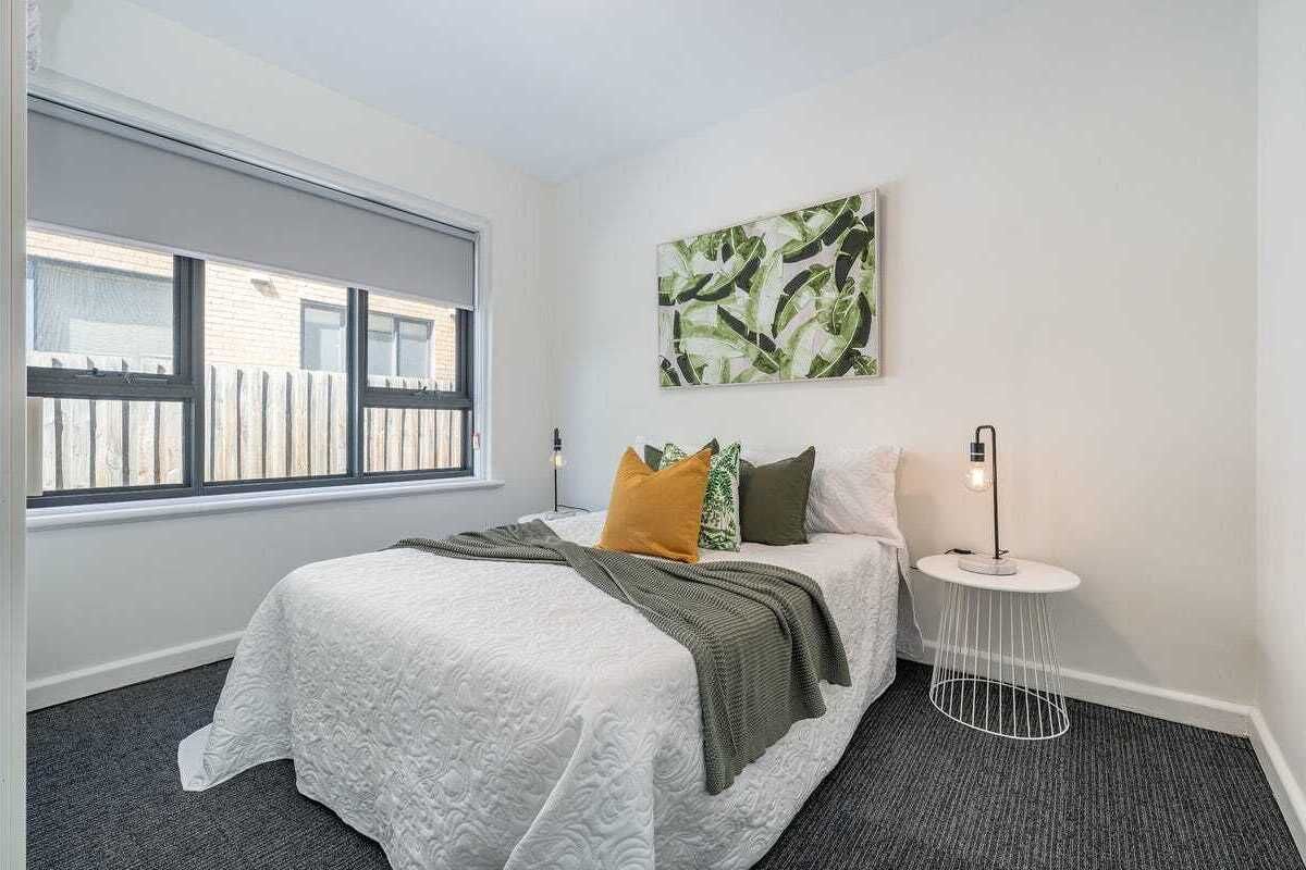 A bedroom with a large window, a bed with a white quilt, green and yellow pillows, a round side table with a lamp, and a wall-mounted abstract painting.