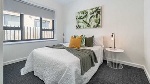 A bedroom with a large window, a bed with a white quilt, green and yellow pillows, a round side table with a lamp, and a wall-mounted abstract painting.