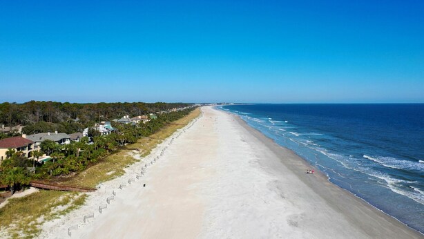 A long, sandy beach with ocean waves, lined with houses and greenery.