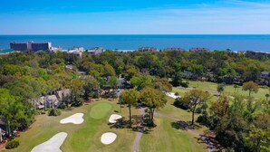 An aerial view of a golf course with sand bunkers, a clear view of the ocean, and surrounding residential buildings.