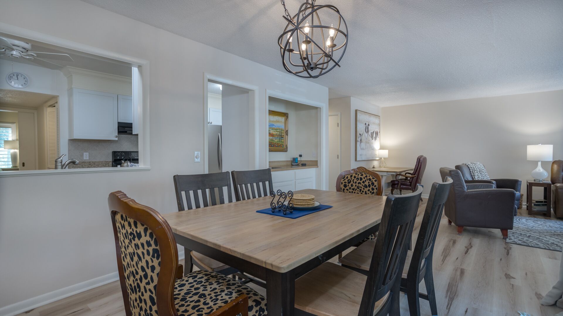A dining area with a wooden table, chairs, and a chandelier. There is a kitchen visible through an open doorway.