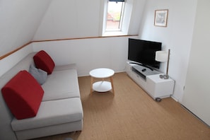 Une petite pièce moderne avec un canapé gris, des coussins rouges, une table blanche, un téléviseur posé sur un meuble blanc et une fenêtre donnant sur un immeuble.