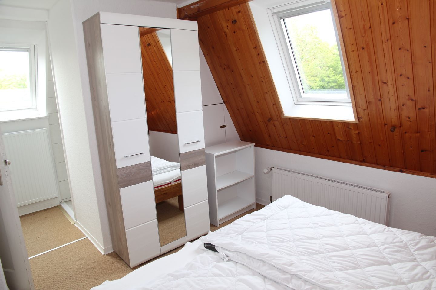 A bedroom with a sloped ceiling, a white wardrobe, a bed with white bedding, and a window with a view of greenery.