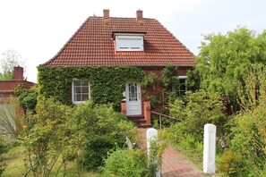 Une maison en briques avec un toit recouvert de tuiles rouges, des murs couverts de lierre et une porte d’entrée blanche.