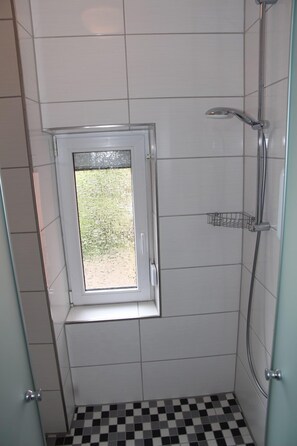 Une petite douche carrelée avec une fenêtre, un sol à carreaux et une étagère en métal.