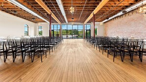 Un grand hall spacieux, avec des rangées de chaises et de tables, un plancher en bois et un plafond élevé avec des poutres apparentes et des conduits visibles.