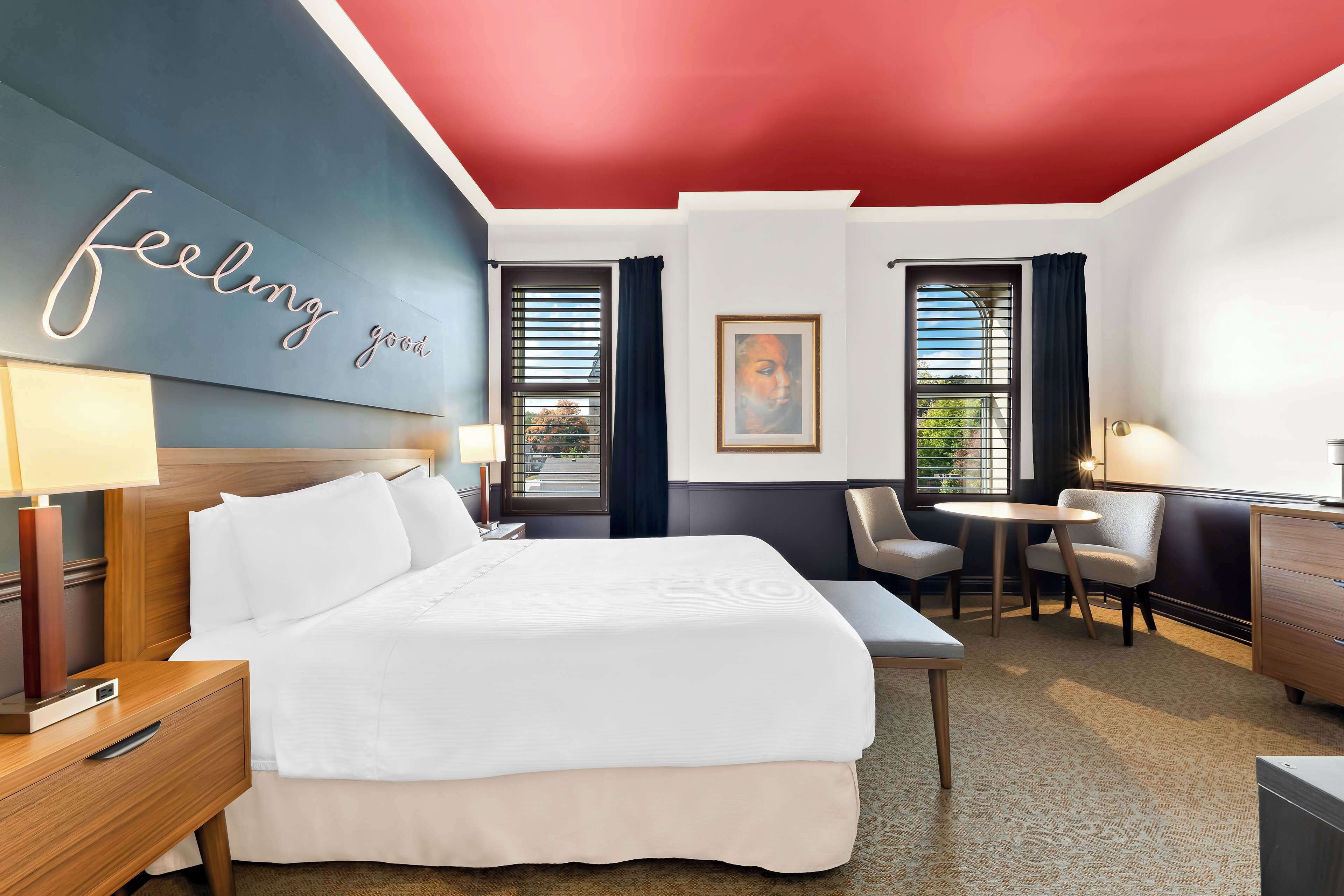 Ein Hotelzimmer mit Bett, Nachttischen, Schreibtisch und Stuhl. Es gibt ein Fenster mit Vorhängen, eine Wand mit einem gerahmten Bild und eine Decke mit rotem Bereich.