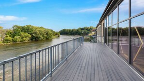Une terrasse en bois avec des rampes en métal, donnant sur une rivière, entourée d’arbres et sous un ciel dégagé.