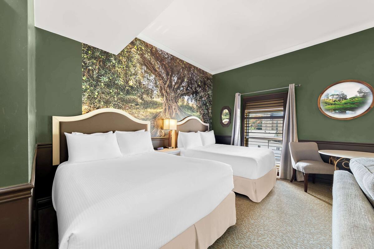 Habitación de hotel con dos camas, un sofá, una mesita y vista a un árbol a través de una ventana.