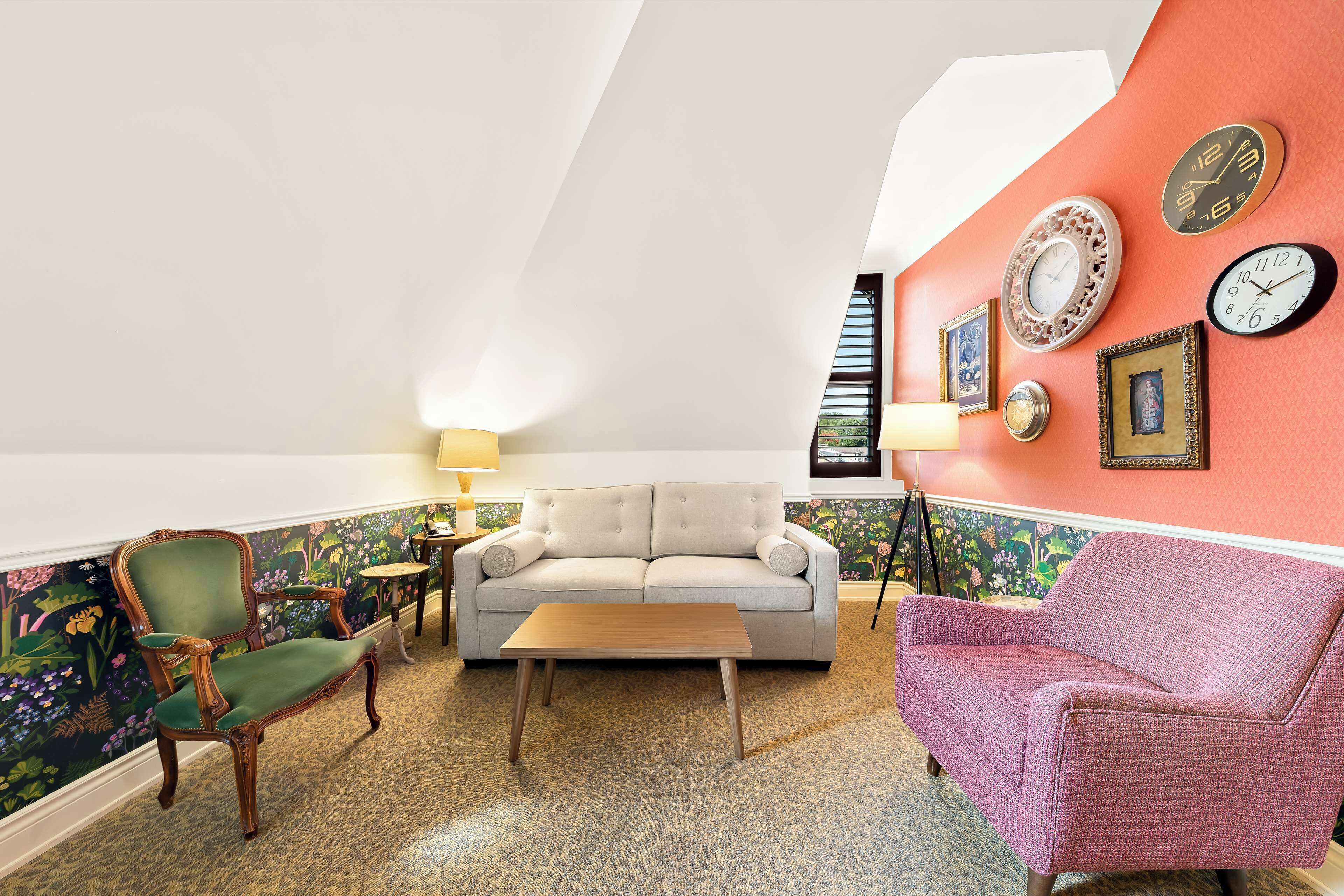 A cozy living room with a green armchair, a beige sofa, and a pink sofa. There are clocks on the wall and a lamp on a side table.