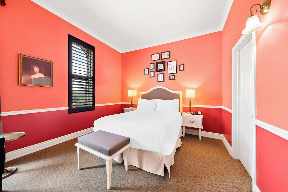 Habitación de hotel con una cama grande, mesitas de noche con lámparas, un banco y una pared con cuadros.