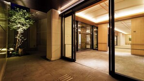 Entrada de un edificio moderno con puertas de cristal, suelo de baldosas y un pequeño árbol en el exterior.