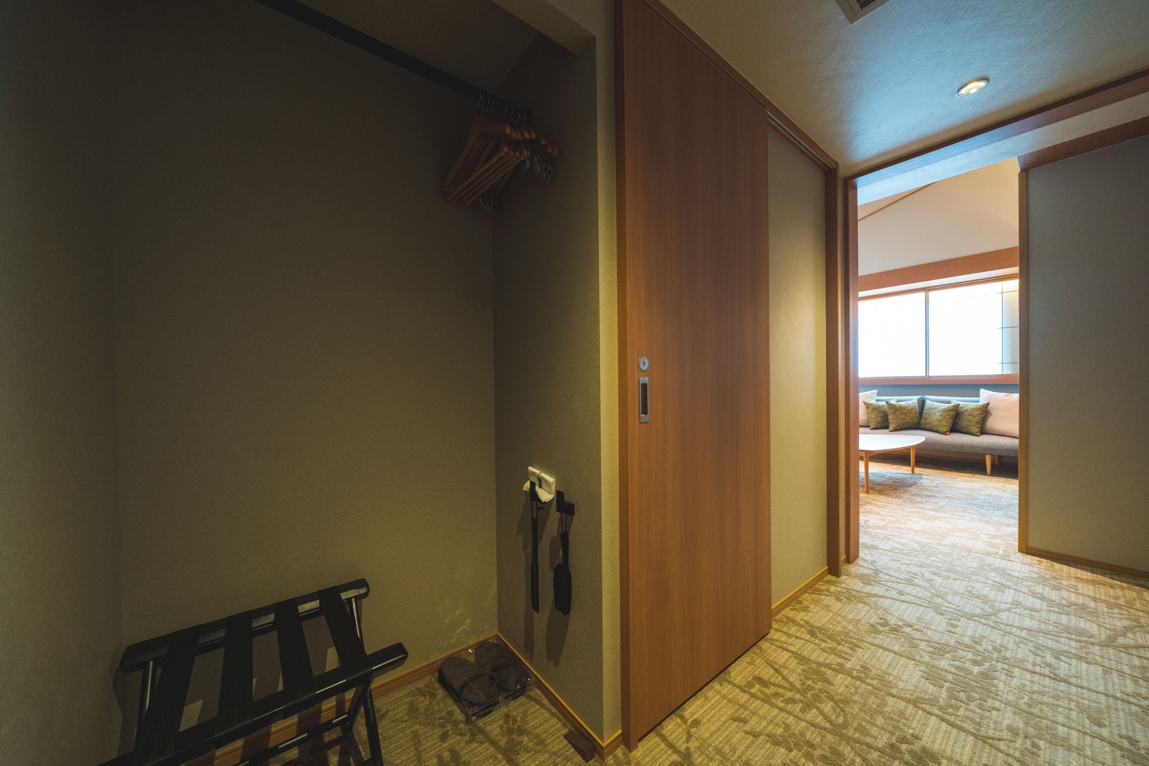 A hotel hallway with a bench, a coat rack, and a door leading to a room with a sofa and a window.
