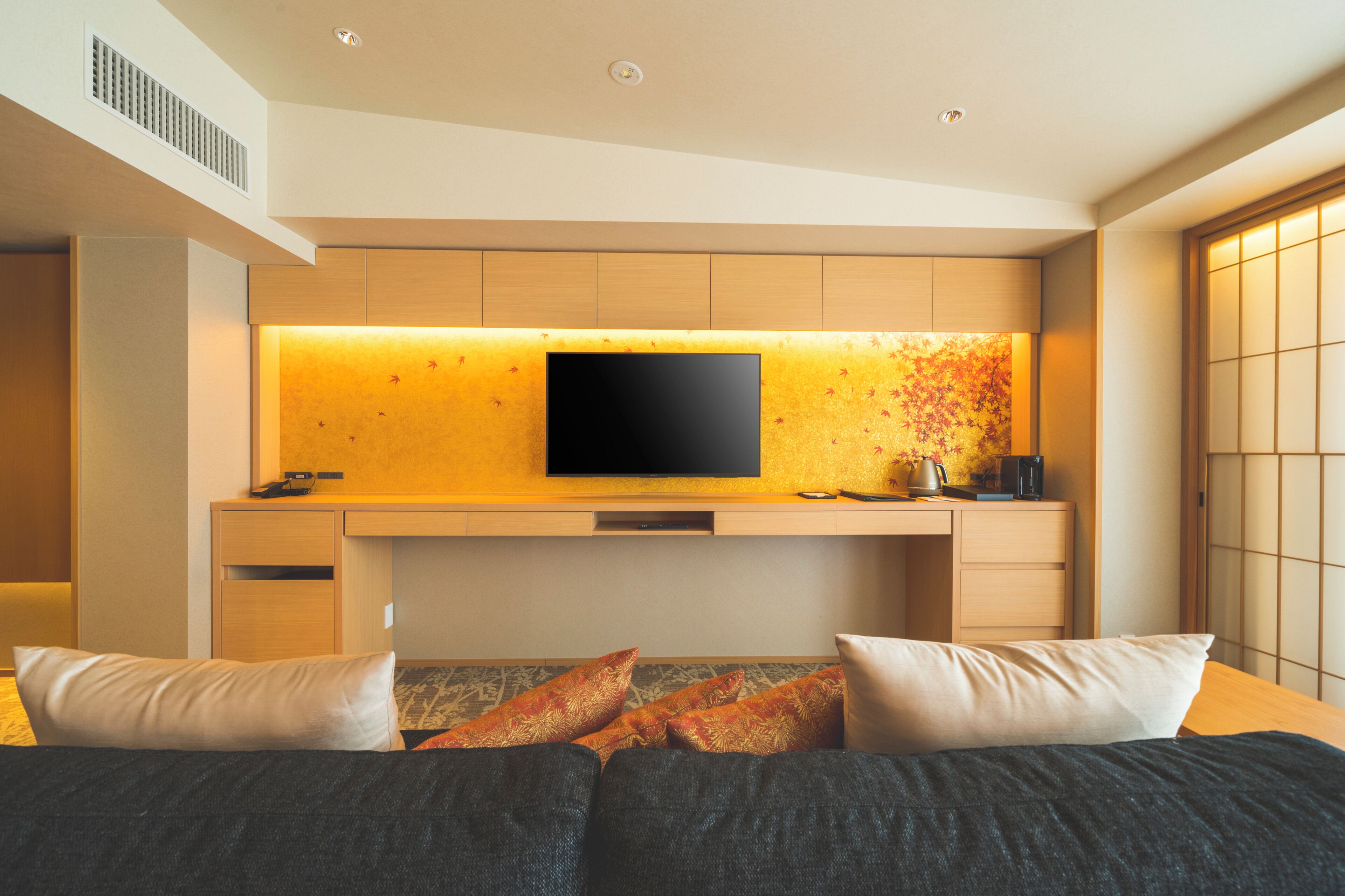 A modern hotel room with a flat-screen TV mounted on a wall, a bed with dark bedding, and a desk with a computer and a lamp.