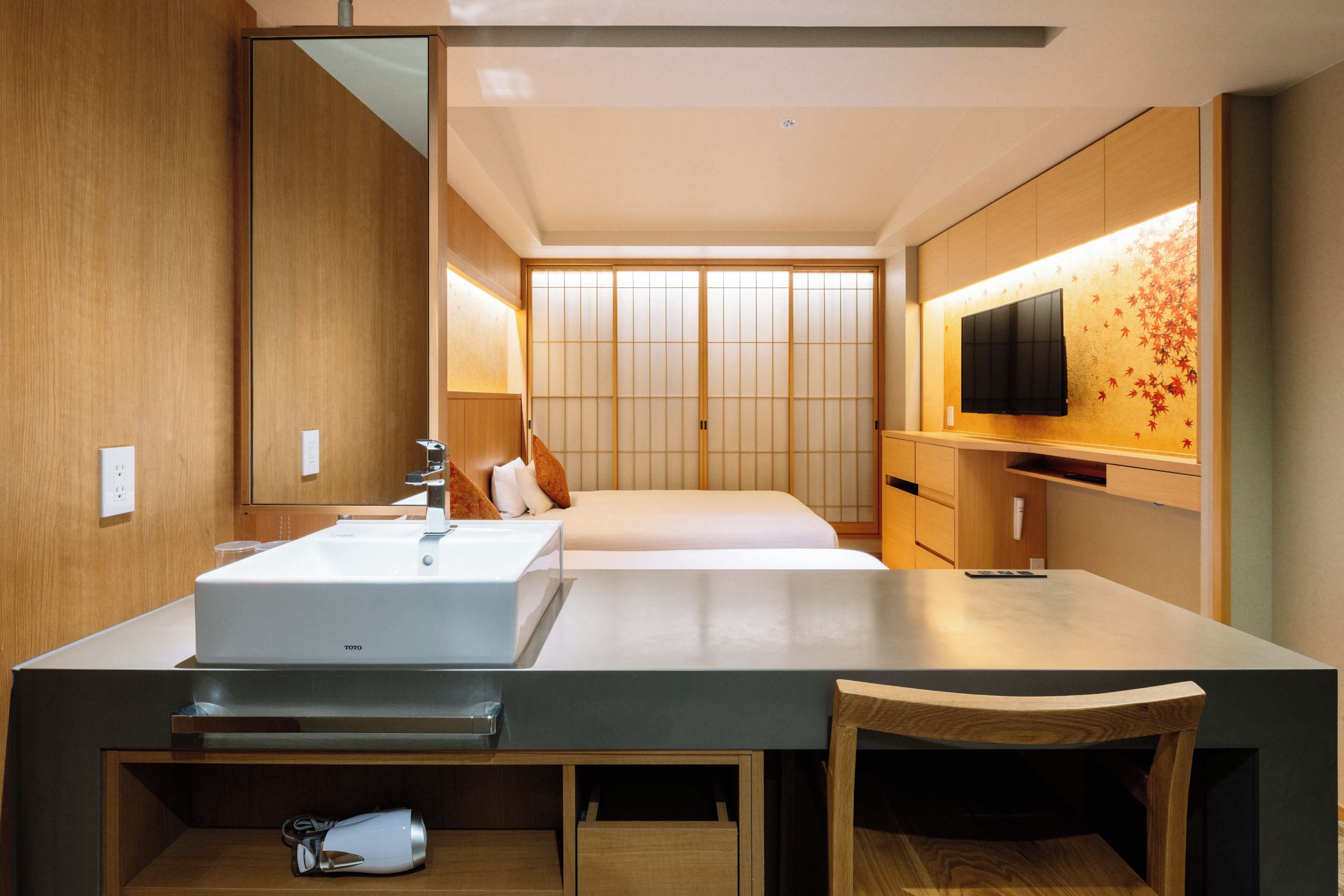 A modern hotel room with a large bed, a bedside table, a mirror, a TV mounted on the wall, and a wooden cabinet.