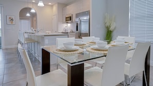 Una cocina moderna con una isla central, electrodomésticos de acero inoxidable y un comedor con mesa de vidrio y sillas blancas.