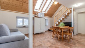 Un espacio habitable compacto con un entrepiso para dormir, una zona de comedor con sillas de madera, un sofá gris y una cocina con un armario blanco y un refrigerador.