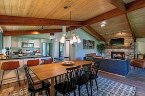 A spacious dining area with a wooden table and chairs, a kitchen in the background, and a stone fireplace.