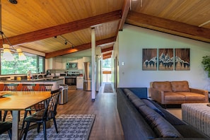 A spacious living area with a wooden ceiling, a dining table with chairs, a sofa, and a kitchen in the background.