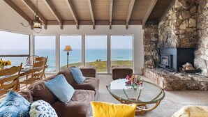 A cozy living room with a stone fireplace, a glass coffee table, and a view of the ocean. - Rustic Oceanfront Cabin (Dillon Beach)