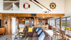 A spacious living area with a brown sofa, a glass coffee table, wooden furniture, and a kitchen in the background. - Rustic Oceanfront Cabin (Dillon Beach)