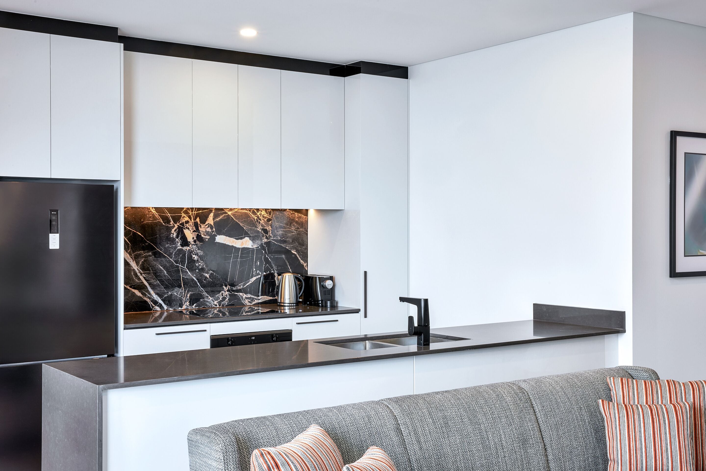 A modern kitchen with white cabinets, a black marble backsplash, and a grey sofa with striped pillows.