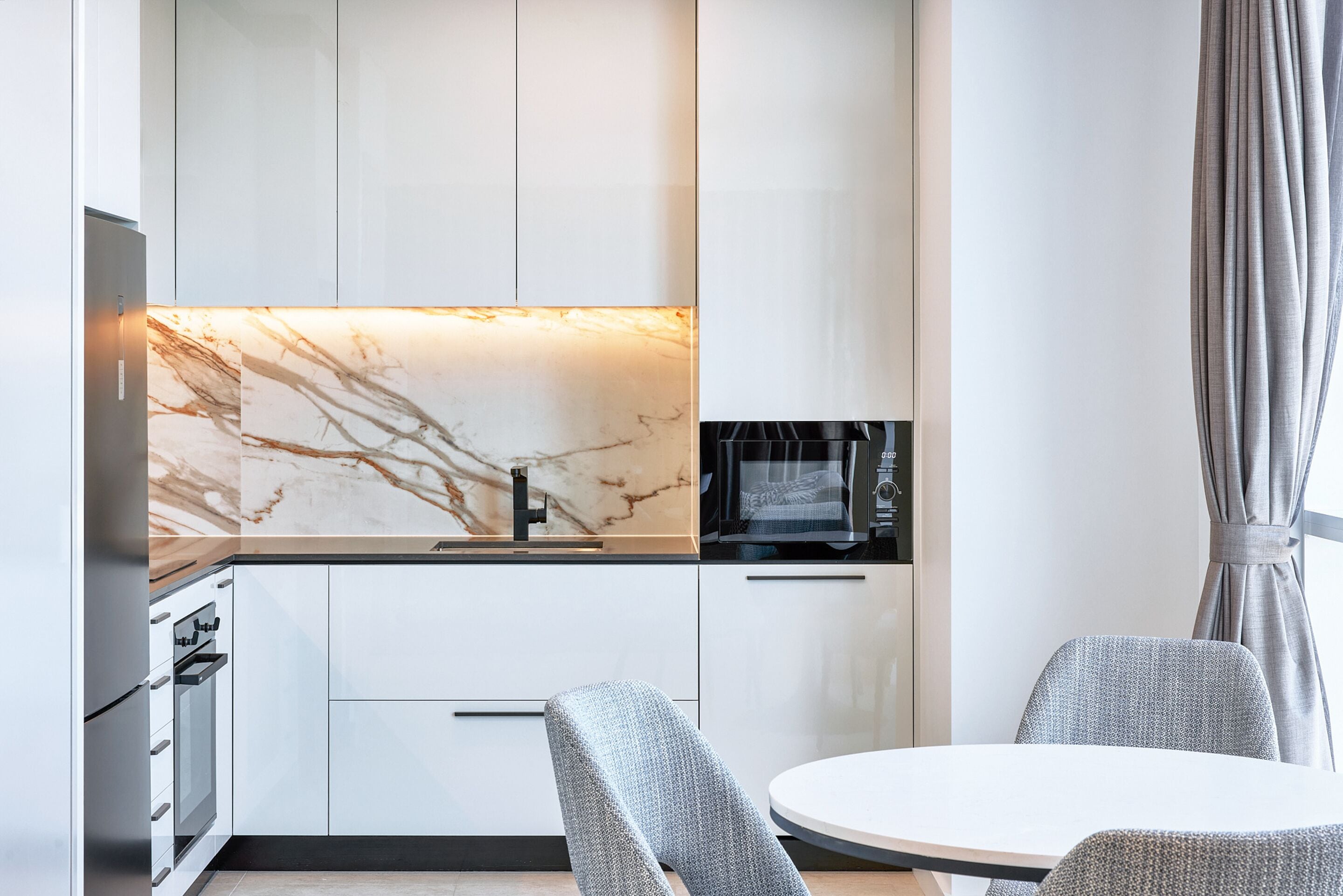 A modern kitchen with white cabinets, a marble backsplash, a built-in oven, and a microwave. There is a round dining table with two chairs.
