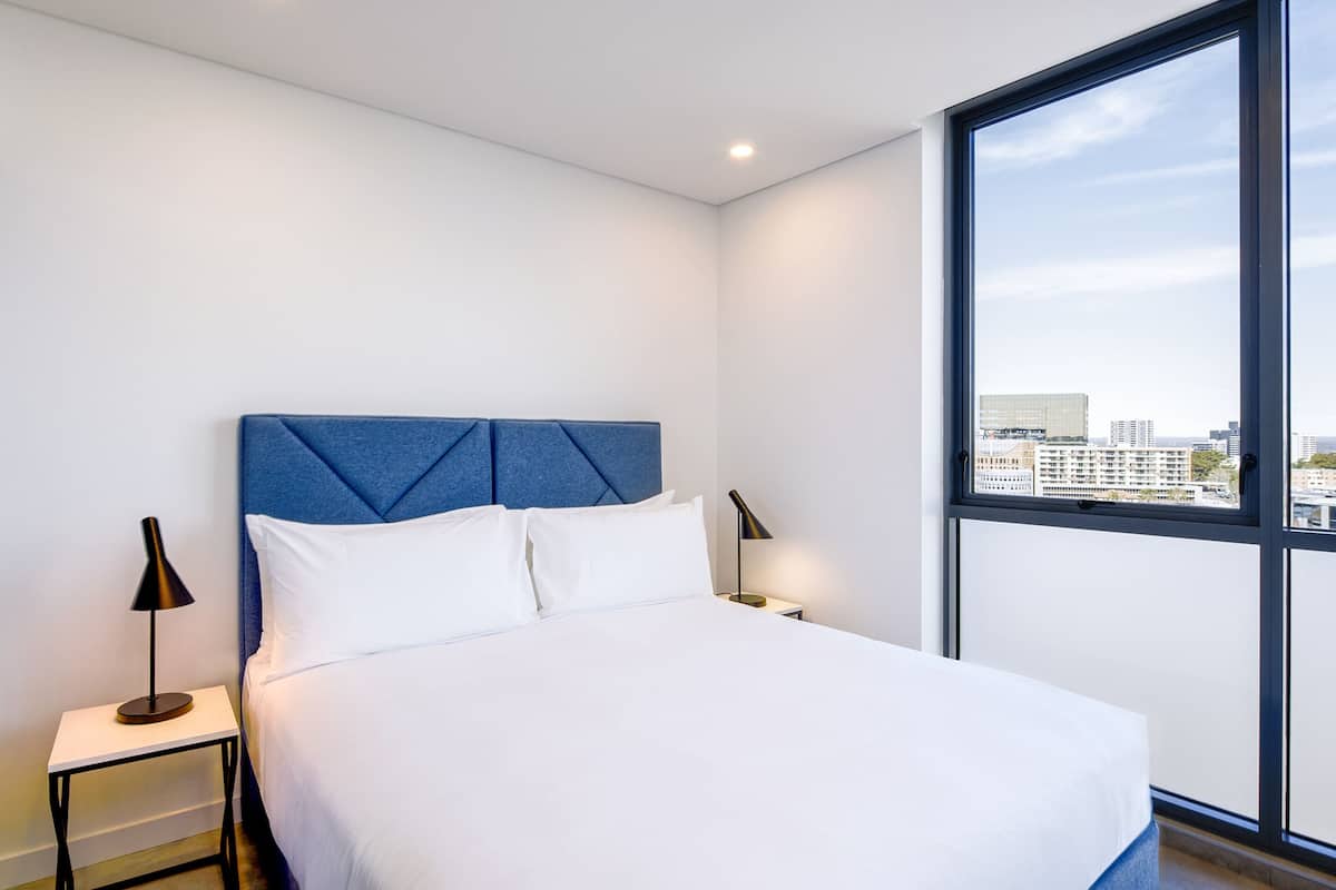 A modern hotel room with a large bed, a blue headboard, white bedding, two bedside tables with lamps, and a large window offering a city view.