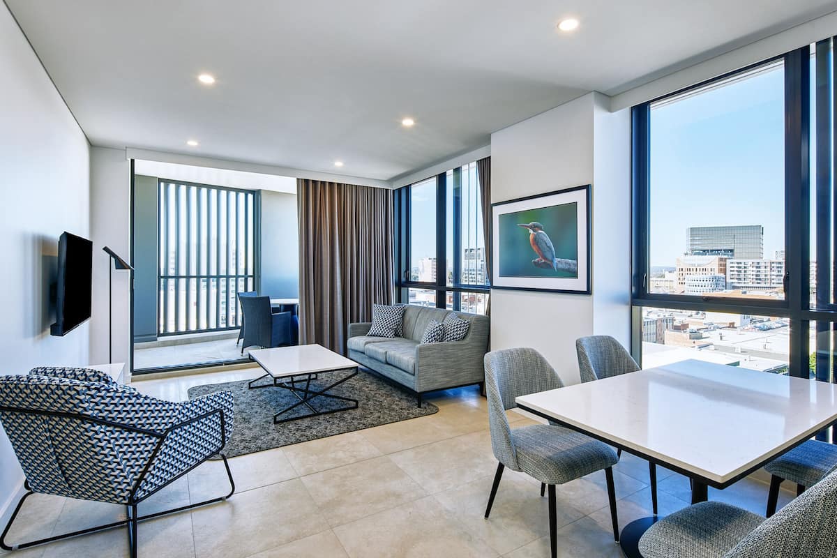 A modern hotel room with a large window, a sofa, a dining table, and a flat-screen TV.