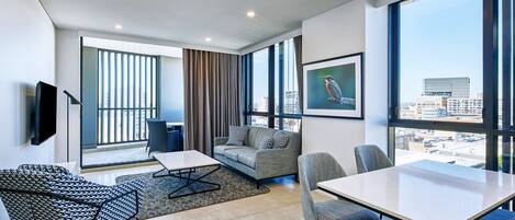 Ein modernes Hotelzimmer mit großem Fenster, einer Couch, einem Esstisch und einem Flachbildfernseher.
