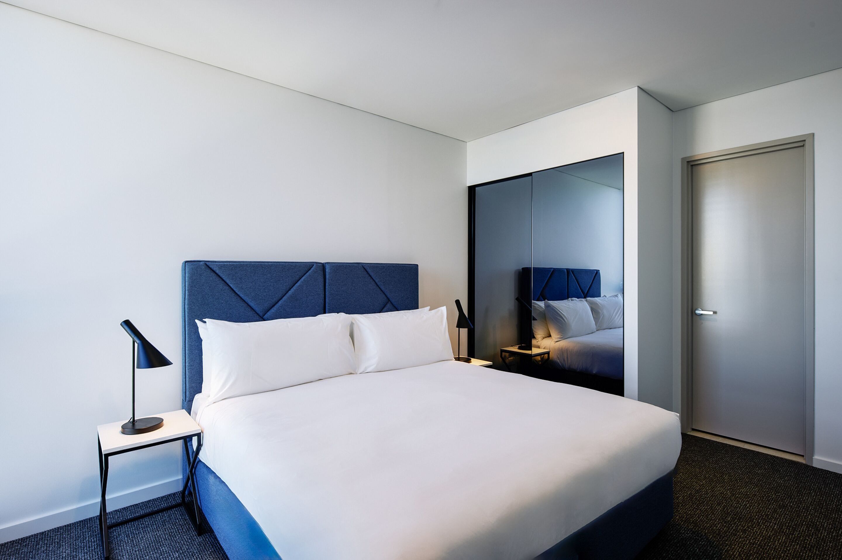A modern hotel room with a large bed, a nightstand, a lamp, and a wardrobe.