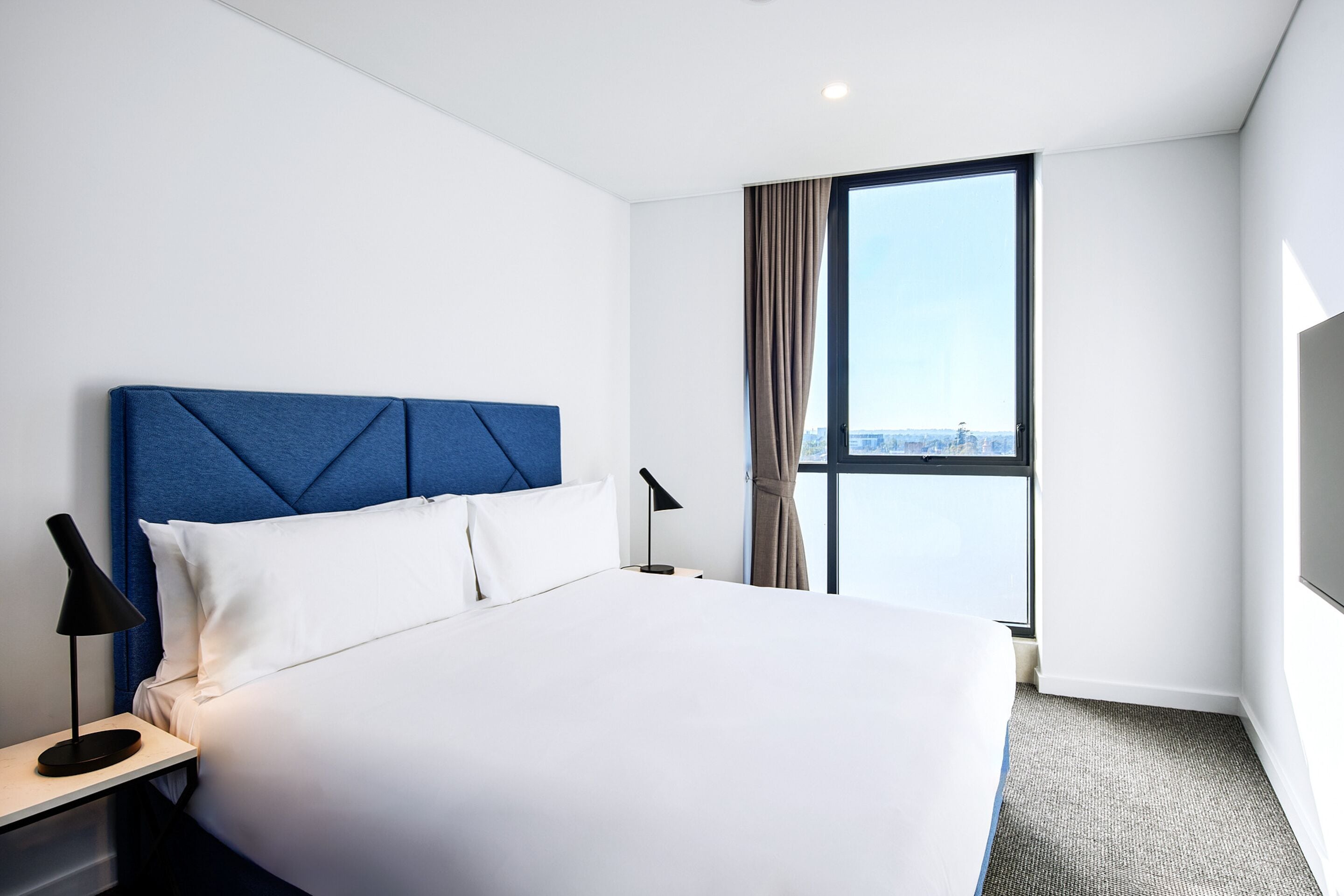 A hotel room with a large bed, a nightstand with a lamp, a window with curtains, and a view of the city.