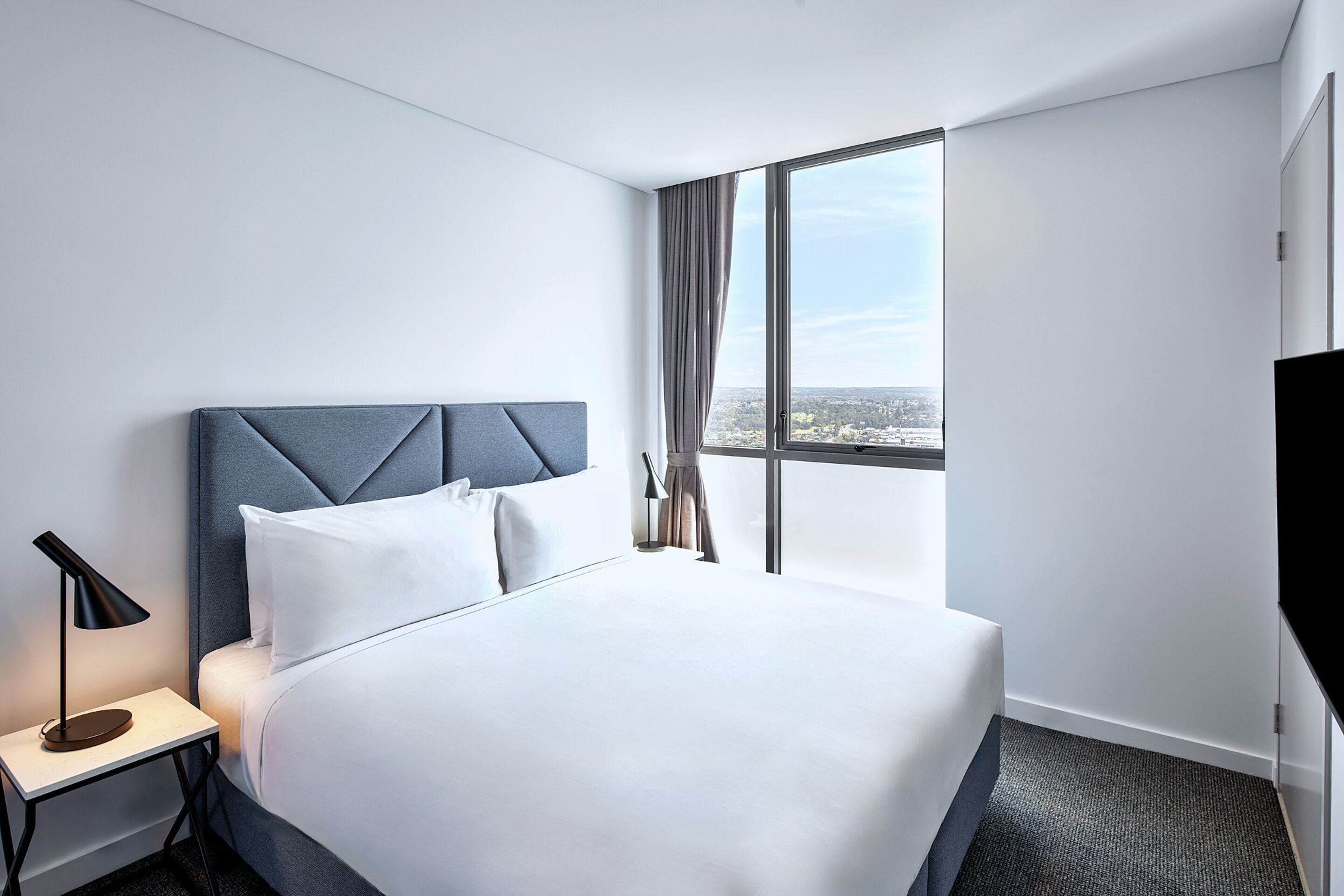 Quarto de hotel com uma cama grande, uma mesinha de cabeceira com abajur, uma janela com cortinas e vista para a cidade.
