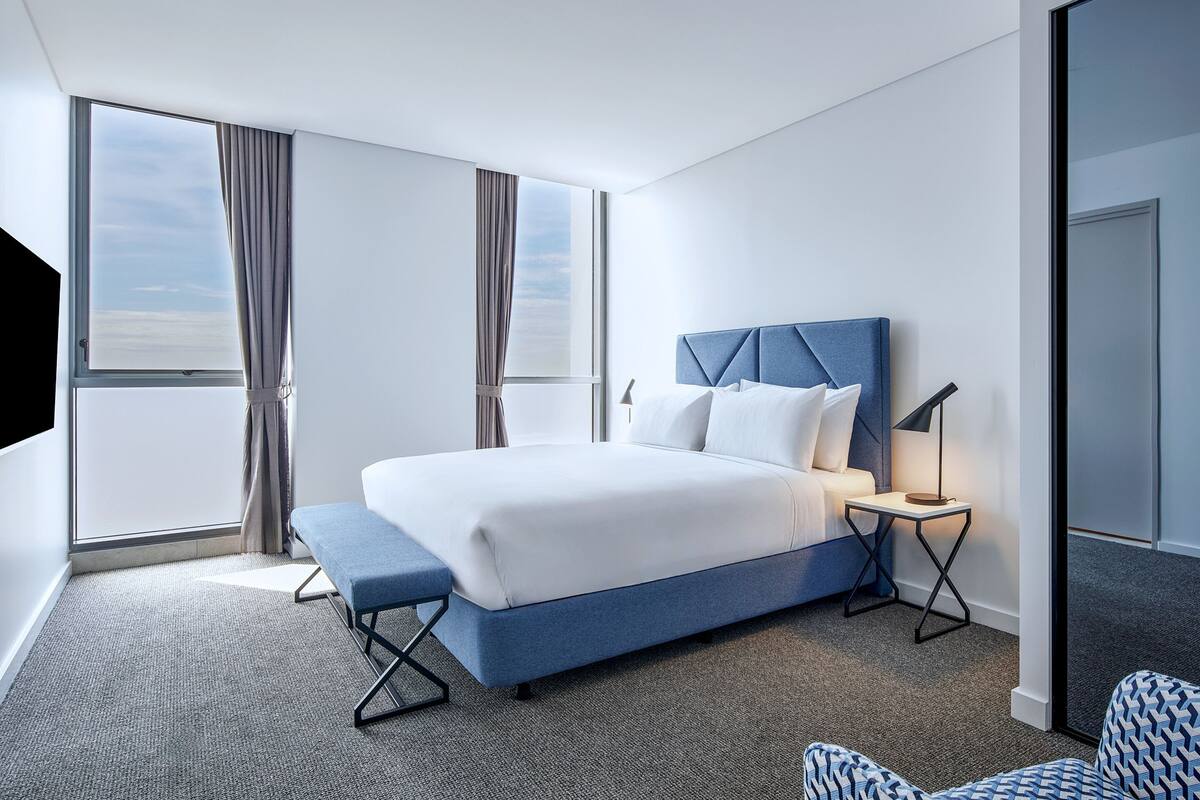 A modern hotel room with a large bed, a bedside table, a bench, and a view of the sky through the window.