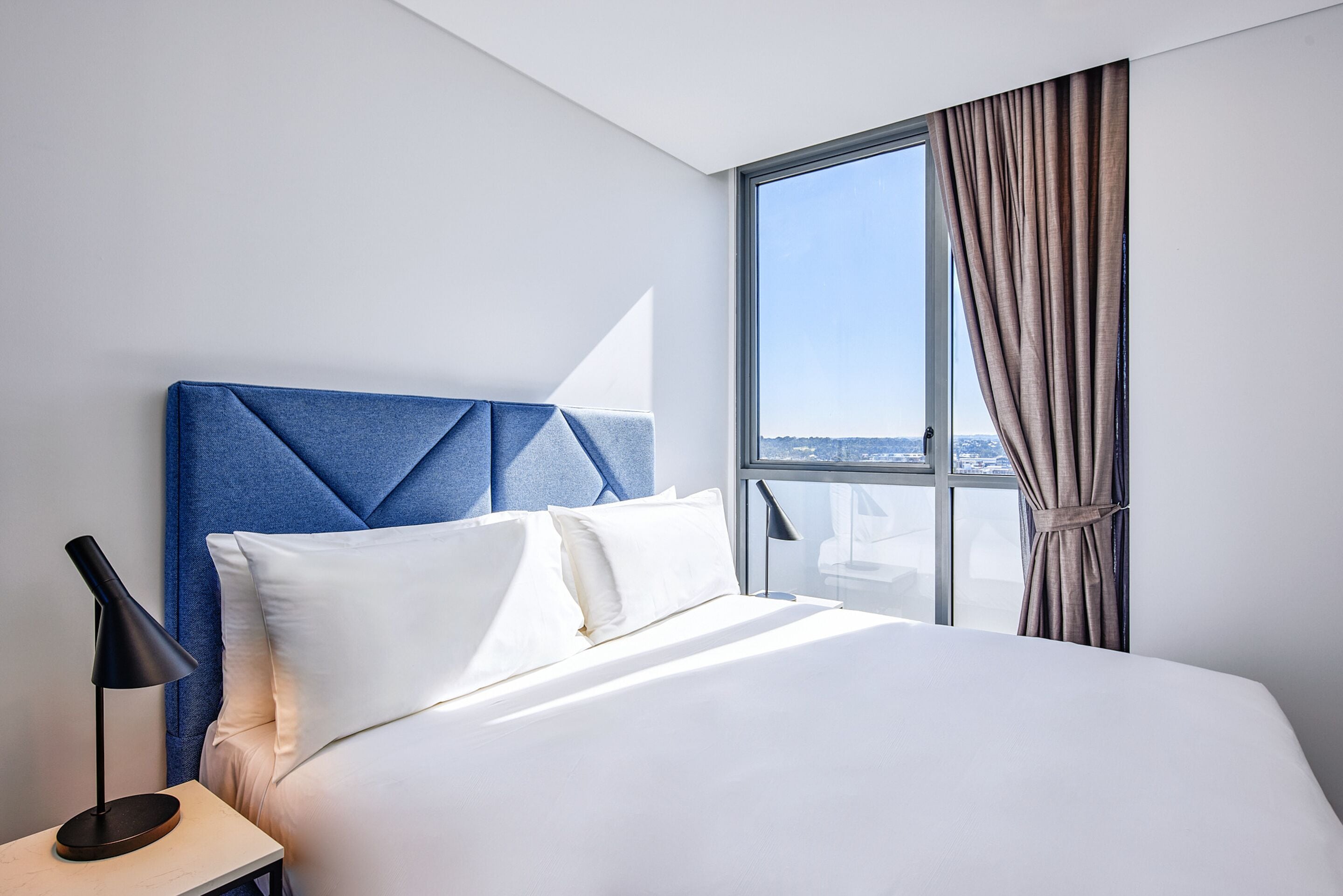 Una habitación de hotel moderna con una cama grande, una mesita de noche, una lámpara y vistas a la ciudad a través de un ventanal amplio.