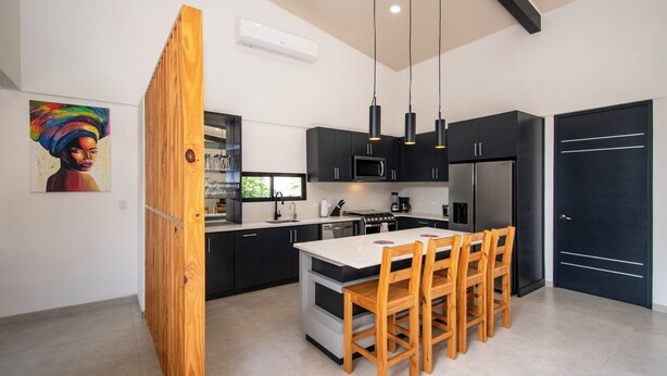 A modern kitchen with dark cabinetry, a central island, wooden chairs, and pendant lighting.