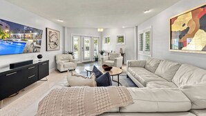 A modern living room with a large flat-screen TV, a beige sectional sofa, and a black entertainment unit. - Mermaids Oasis | Private Pool, Hot Tub, & 5 Minute Walk to St. Armands Circle & Lido Key Beach! (Lido Key)